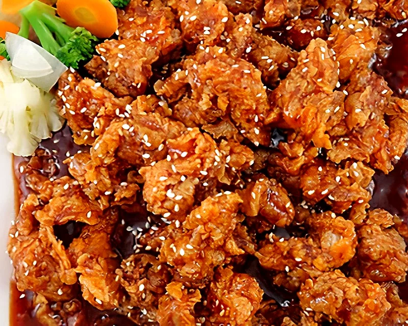 Korean fried chicken pieces in a glossy sweet chili glaze with sesame seeds and steamed vegetables on a square plate